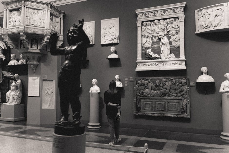 A grayscale photo of a museum exhibit featuring classical sculptures and a visitor observing the artworks.